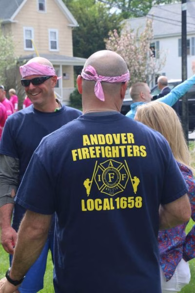 A group of people outdoors, including a person wearing a shirt that reads Andover Firefighters Local 1658 and a pink bandana. Others nearby also wear blue shirts and pink bandanas. Houses are visible in the background.