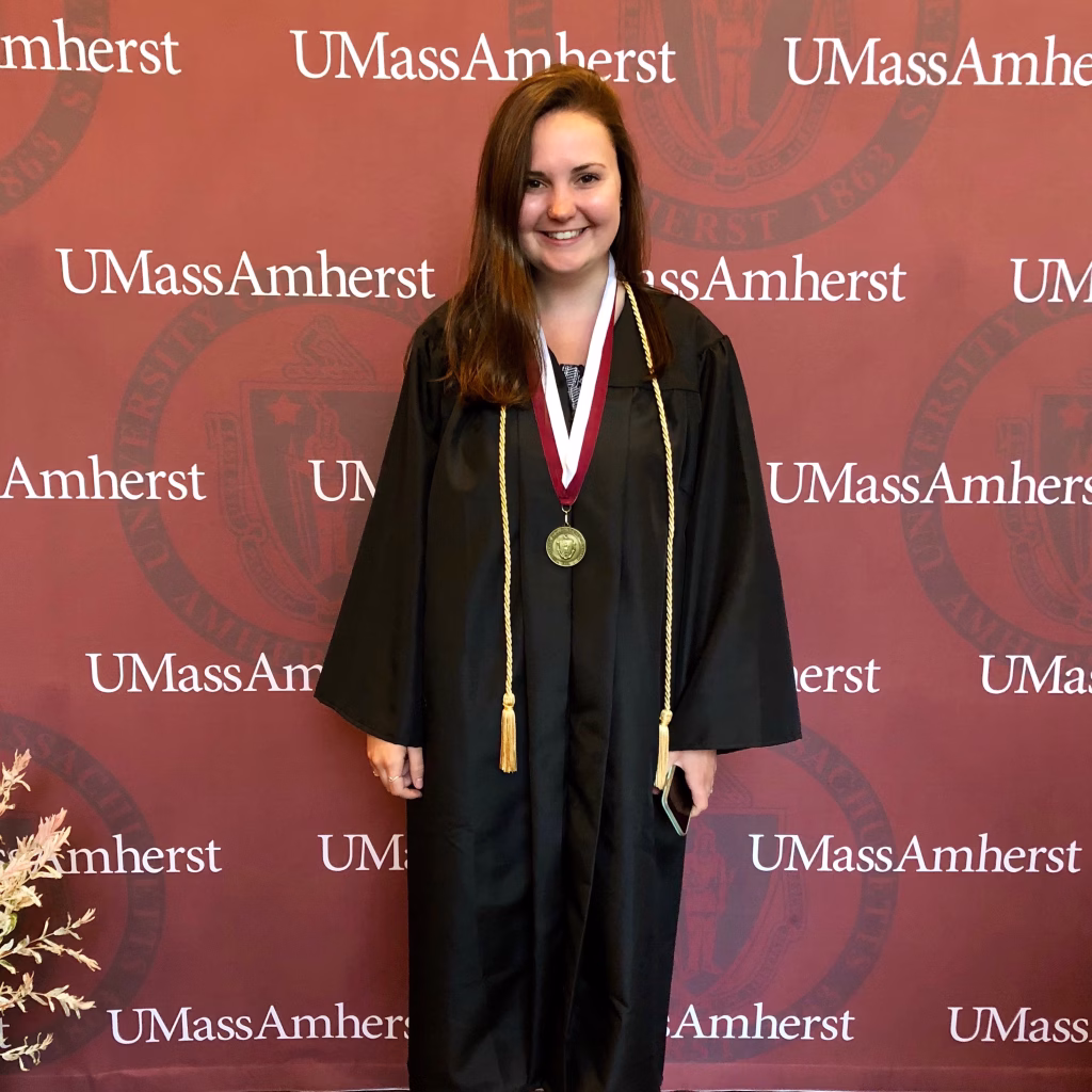 A young woman wearing a graduation gown and medal stands in front of a UMass Amherst backdrop, smiling at the camera.