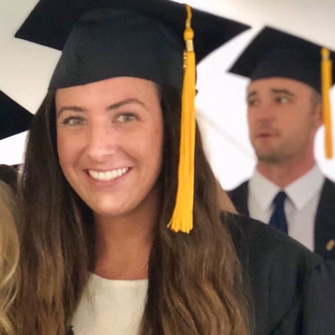 A smiling graduate wearing a black cap with a yellow tassel and gown stands in the foreground, with another graduate in similar attire blurred in the background.