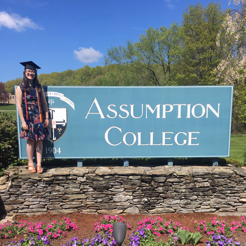 A young woman in a graduation cap and dress stands beside a large Assumption College sign, surrounded by flowers, with trees and blue sky in the background.