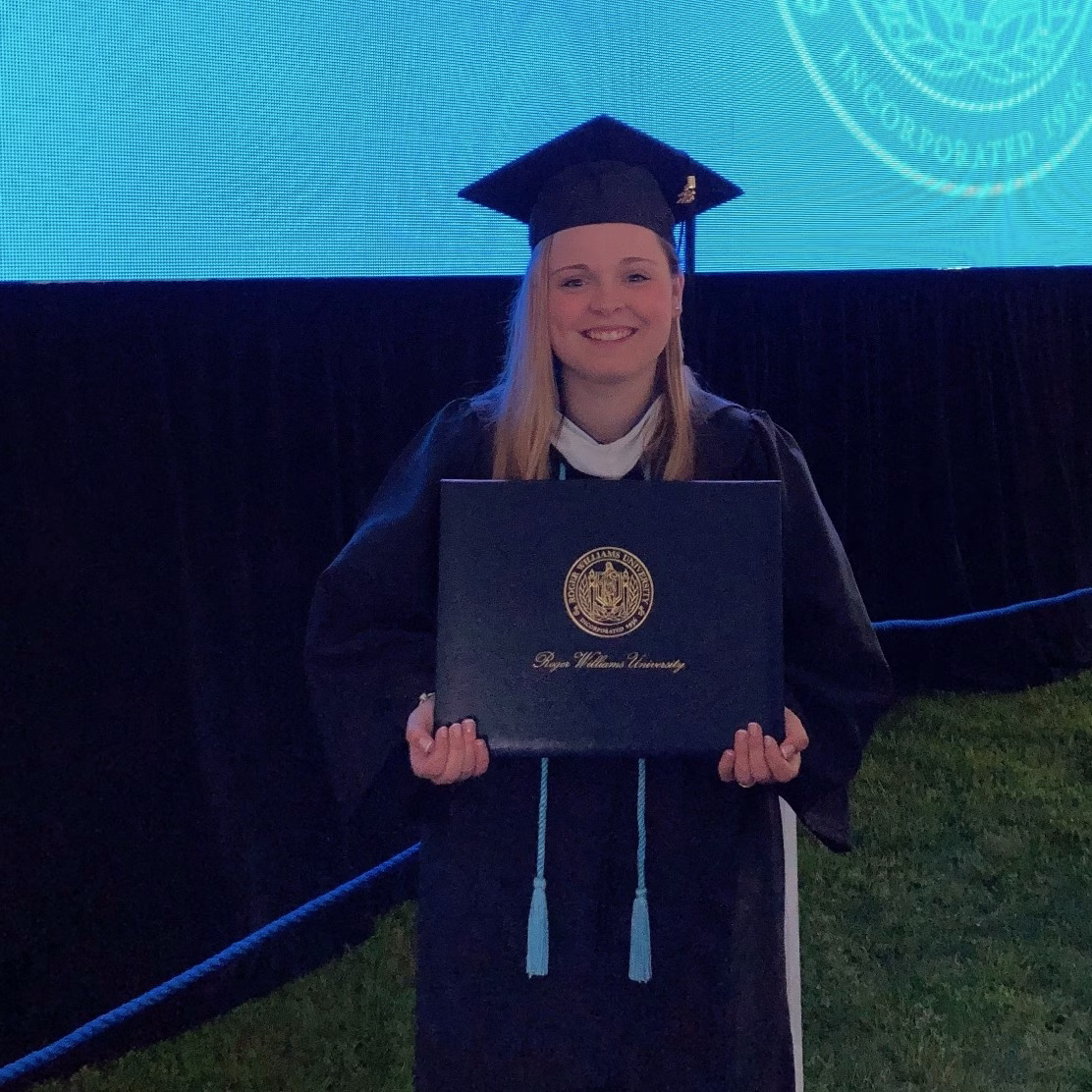 A graduate in a cap and gown smiles while holding a diploma cover in front of a large blue university backdrop on grass.