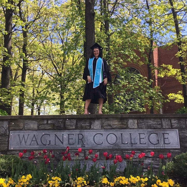A graduate in cap and gown stands on a stone sign that reads “Wagner College,” surrounded by blooming red and yellow flowers and green trees on a sunny day.