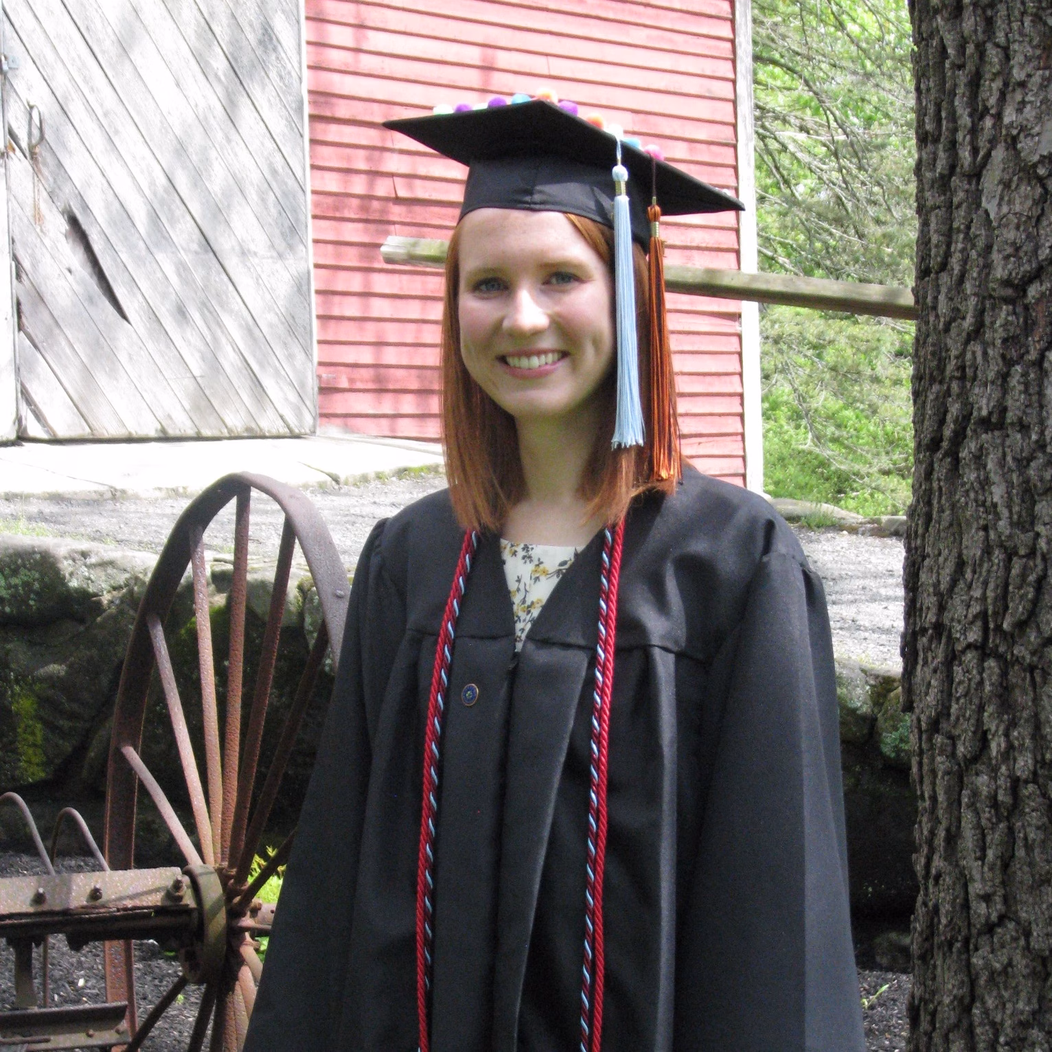 A young woman in a black graduation gown and cap smiles outdoors beside a tree. An old wooden wheel and a rustic red building are visible in the background.