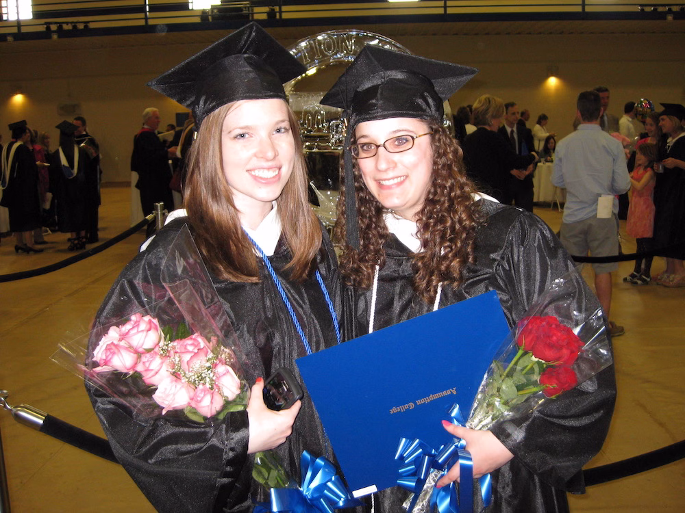 Two graduates in black caps and gowns smile at the camera, each holding flowers and a diploma. People in graduation attire are gathered in the background inside a large hall.
