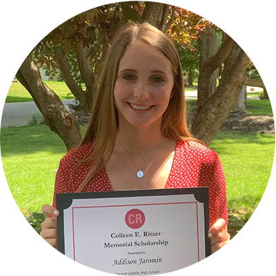 A young woman with long hair, wearing a red polka-dot blouse, smiles outdoors while holding a framed certificate for the Colleen E. Ritzer Memorial Scholarship. Trees and greenery are in the background.