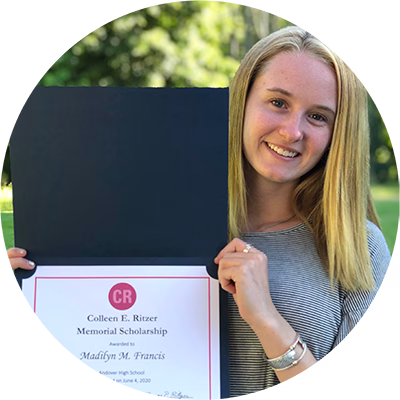 A young woman with straight blonde hair smiles while holding up a scholarship certificate outdoors. The certificate is labeled Colleen E. Ritzer Memorial Scholarship and awarded to Madilyn M. Francis.