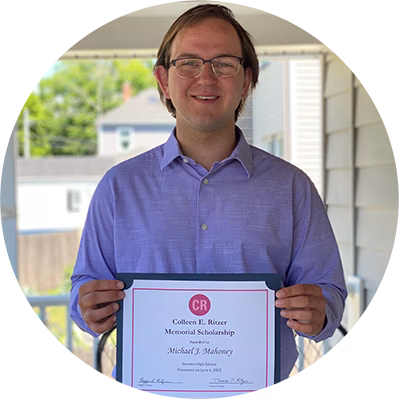 A young man in glasses and a light purple shirt smiles while holding a Colleen E. Ritzer Memorial Scholarship certificate on a porch with a blurred house and greenery in the background.
