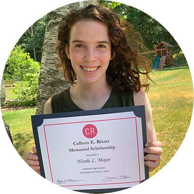 A young woman with curly brown hair smiles while holding a Colleen E. Ritzer Memorial Scholarship certificate outdoors, with trees and a playground in the background.
