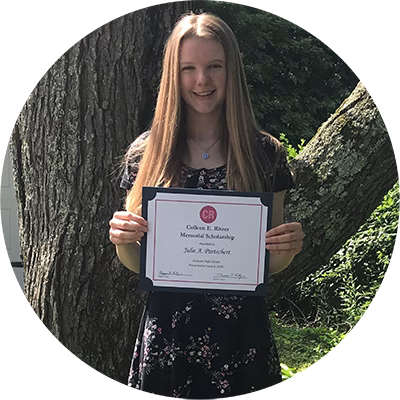 A young woman with long, straight hair stands outside in front of a tree, smiling and holding a scholarship certificate. She is wearing a floral dress.