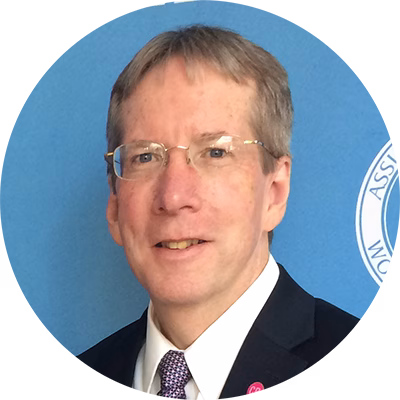 A man with short light brown hair and glasses, wearing a dark suit, white shirt, and patterned tie, poses in front of a blue background with a partial circular logo visible.