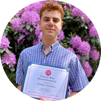 A young man with short red hair, wearing a blue plaid shirt, stands in front of blooming purple flowers while holding a scholarship certificate and smiling slightly.