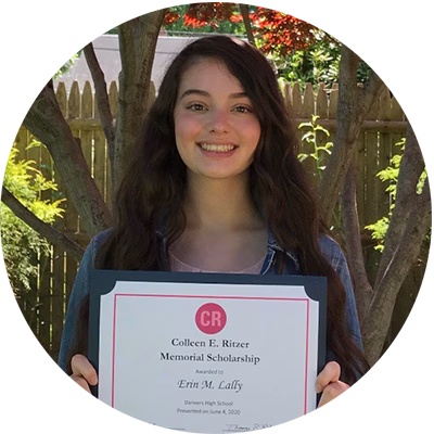 A young woman with long brown hair smiles outdoors, holding a framed certificate for the Colleen E. Ritzer Memorial Scholarship. Trees and a wooden fence are visible in the background.