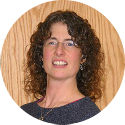 A woman with curly brown hair and glasses is smiling slightly. She is wearing a dark top and standing in front of a wooden background.