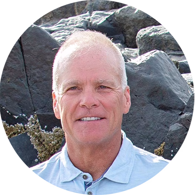A middle-aged man with short light hair, wearing a light blue collared shirt, smiles at the camera. He is outdoors, with large dark rocks in the background.
