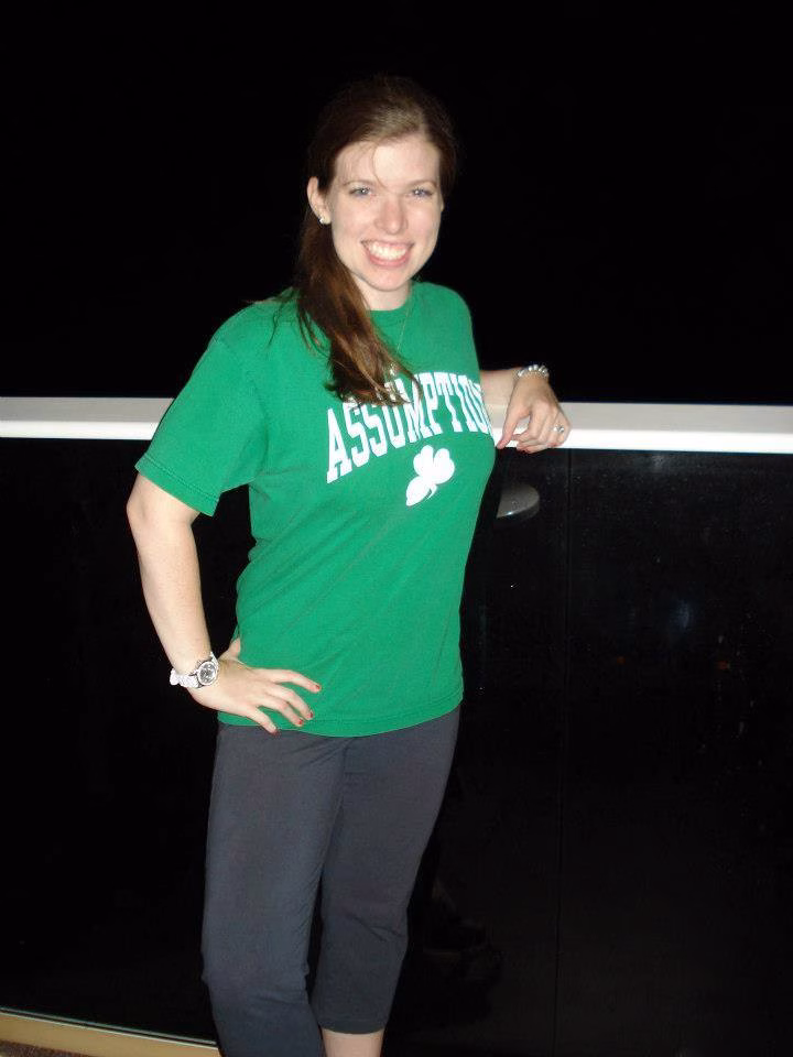 A woman wearing a green ASSUMPTION t-shirt and dark pants stands and smiles in front of a black background and a glass railing.