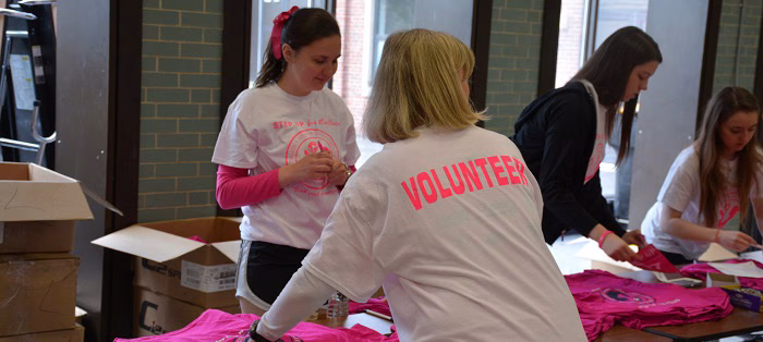 Four women sort and fold bright pink t-shirts at a table indoors. One woman’s shirt says “Volunteer” on the back. Cardboard boxes are nearby, and sunlight comes through large windows.
