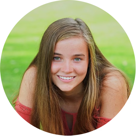 A young woman with long, straight light brown hair smiles at the camera while leaning forward on grass, wearing a coral off-shoulder top. The background is a blurred, bright green lawn.
