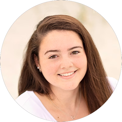 A young woman with long brown hair smiles at the camera. She is wearing a white top and a small necklace, with a blurred, light-colored background.