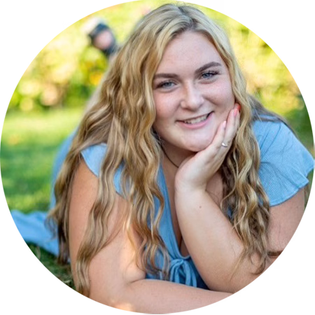 Young woman with long wavy blonde hair, wearing a light blue top, lies on the grass outdoors, smiling at the camera with her hand resting under her chin. The background is blurred greenery.
