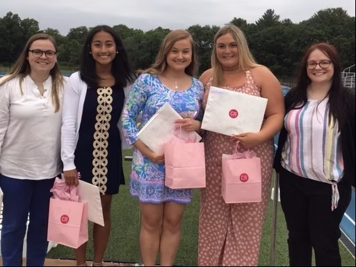 Five young women stand outdoors on a sports field, smiling and holding gift bags and envelopes. They are dressed in semi-formal clothing, and trees are visible in the background under a cloudy sky.
