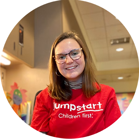 A smiling woman with glasses and shoulder-length brown hair wears a red Jumpstart: Children first. shirt, sitting indoors in a brightly lit room with colorful decorations in the background.