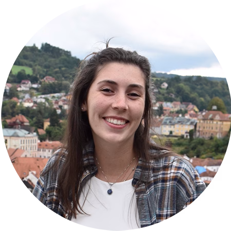 A young woman with long brown hair smiles at the camera, wearing a plaid shirt over a white top. In the background, there is a scenic view of a town with colorful houses and green hills.