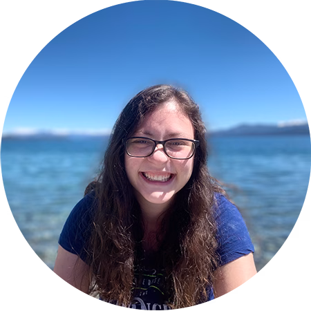 A young woman with long brown hair and glasses smiles at the camera while standing in front of a clear blue lake under a bright sky.