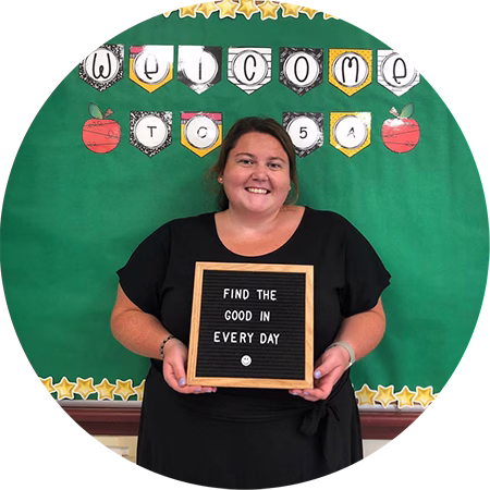 A smiling woman in a black dress holds a letter board that reads Find the good in every day in front of a green bulletin board decorated with the word Welcome and colorful paper apples and stars.