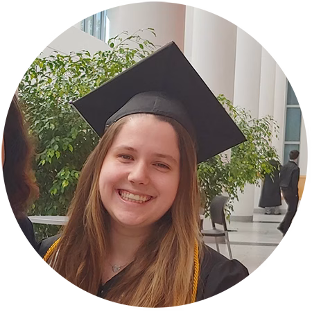 A young woman in a graduation cap and gown smiles indoors, standing in front of green plants and tall white columns.