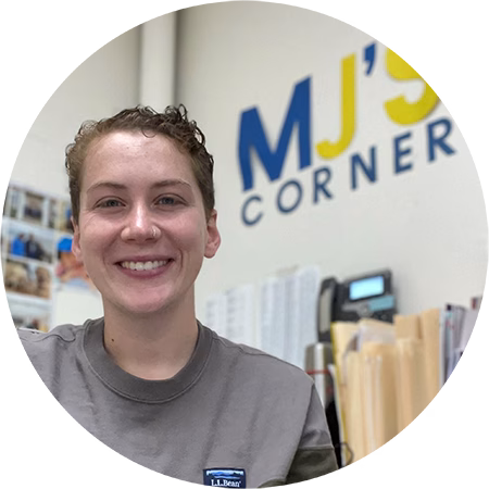 A person with short curly hair wearing a gray shirt smiles at the camera, sitting in an office with MJS CORNER written on the wall behind them and office supplies in the background.