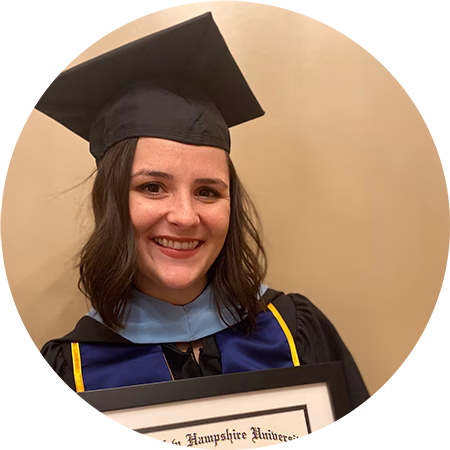 A woman wearing a graduation cap and gown smiles while holding a framed diploma in front of a beige background.