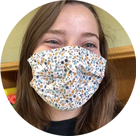 A person with long brown hair smiles while wearing a white face mask with a small, colorful floral pattern. A wooden shelf is visible in the background.