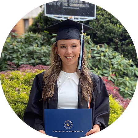 A young woman in a graduation cap and gown smiles while holding a blue Salem State University diploma cover, standing outdoors in front of greenery.