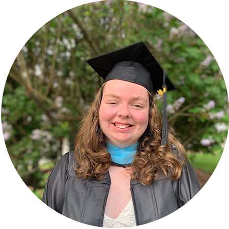 A smiling person with long, curly hair wears a black graduation cap and gown, standing outdoors in front of green trees and flowers.