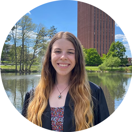 A young woman with long, wavy hair smiles outdoors in front of a pond, wearing a black graduation gown. Trees and a tall brick building are visible in the background under a blue sky.