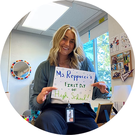A smiling woman with blonde hair sits on a desk holding a sign that reads Ms. Reppuccis First Day of High School! The background shows a classroom with a window, photos, and decorations.