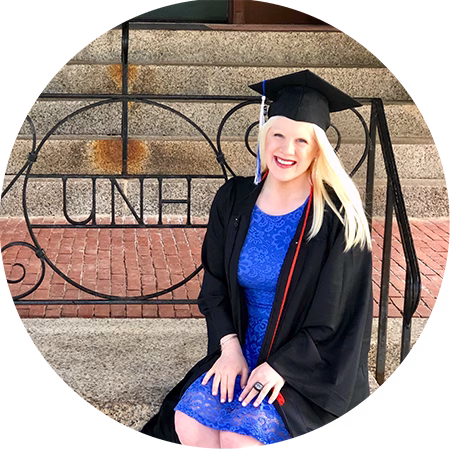 A smiling graduate in a black cap and gown with a blue dress sits on steps in front of a decorative iron railing that spells UNH.