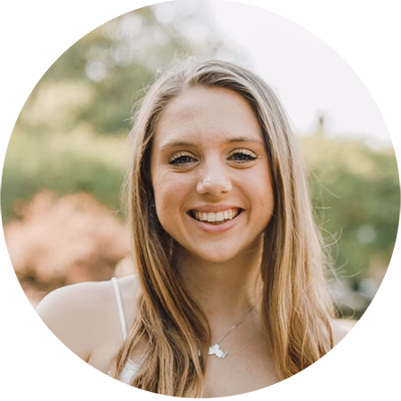 A young woman with long, light brown hair smiles outdoors. She wears a light-colored top and a necklace, with a blurred background of greenery and soft sunlight.