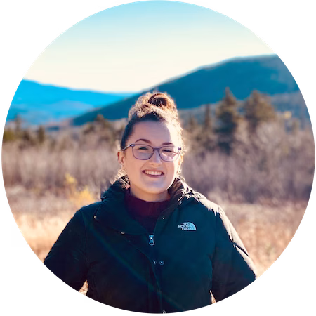 A woman wearing glasses and a dark North Face jacket smiles outdoors, with blurred mountains, trees, and blue sky in the background on a sunny day.