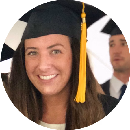 A young woman wearing a graduation cap with a yellow tassel smiles at the camera. Blurred graduates in caps and gowns are visible in the background.