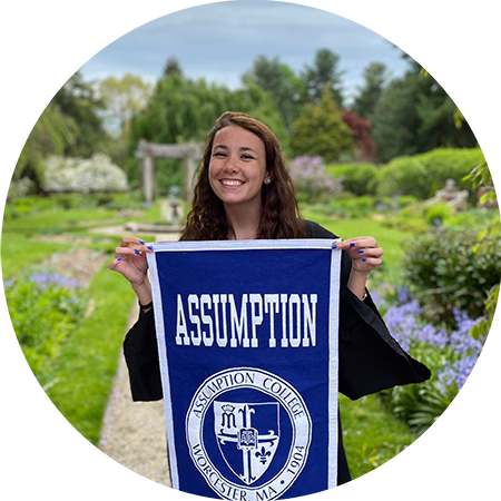 A smiling young woman stands outdoors in a garden, holding a blue and white Assumption College banner. The background features greenery, flowers, and a stone path.