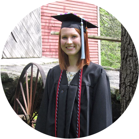A young woman in a black graduation cap and gown stands outside, smiling. A red building, an old wooden wagon wheel, and greenery are visible in the background.