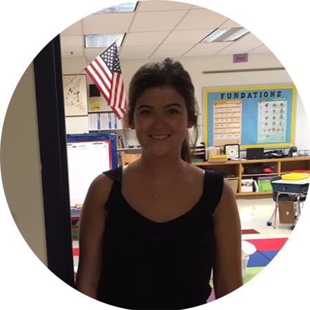 A woman with brown hair wearing a sleeveless black top stands and smiles in a classroom with colorful rugs, an American flag, and educational posters on the wall behind her.