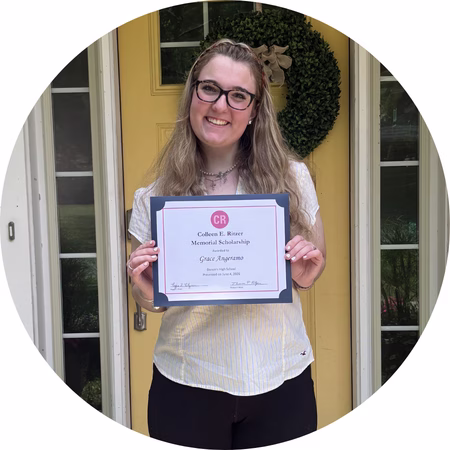 A smiling woman with glasses stands in front of a yellow door, holding a framed certificate or award and wearing a light-colored blouse and dark pants.