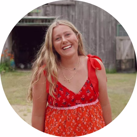 A young woman with long blonde hair smiles outdoors, wearing a red floral dress with ribbon straps and layered necklaces. The background features a rustic wooden building and grass.