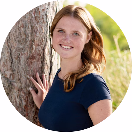 A young woman with light brown hair and a navy blue shirt smiles while standing next to a tree outdoors, with greenery and sunlight in the background.