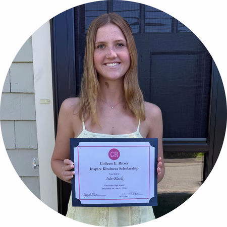 A young woman with long, light brown hair stands outside in front of a door, smiling and holding a certificate for the Colleen R. Ritzer Inspire Kindness Scholarship awarded to Ida Walsh.