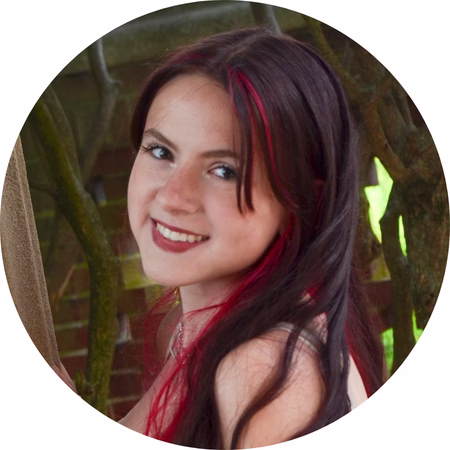 A young woman with long dark hair and red streaks smiles at the camera. She is outdoors, with green foliage and brickwork visible in the background.
