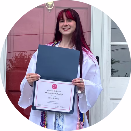 A young woman in a white graduation gown smiles while holding an open folder displaying a scholarship certificate. She stands in front of a red door.