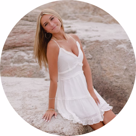 A young woman with long blonde hair, wearing a sleeveless white dress, sits on a large rock outdoors and smiles at the camera. The background features more rocks, creating a natural setting.